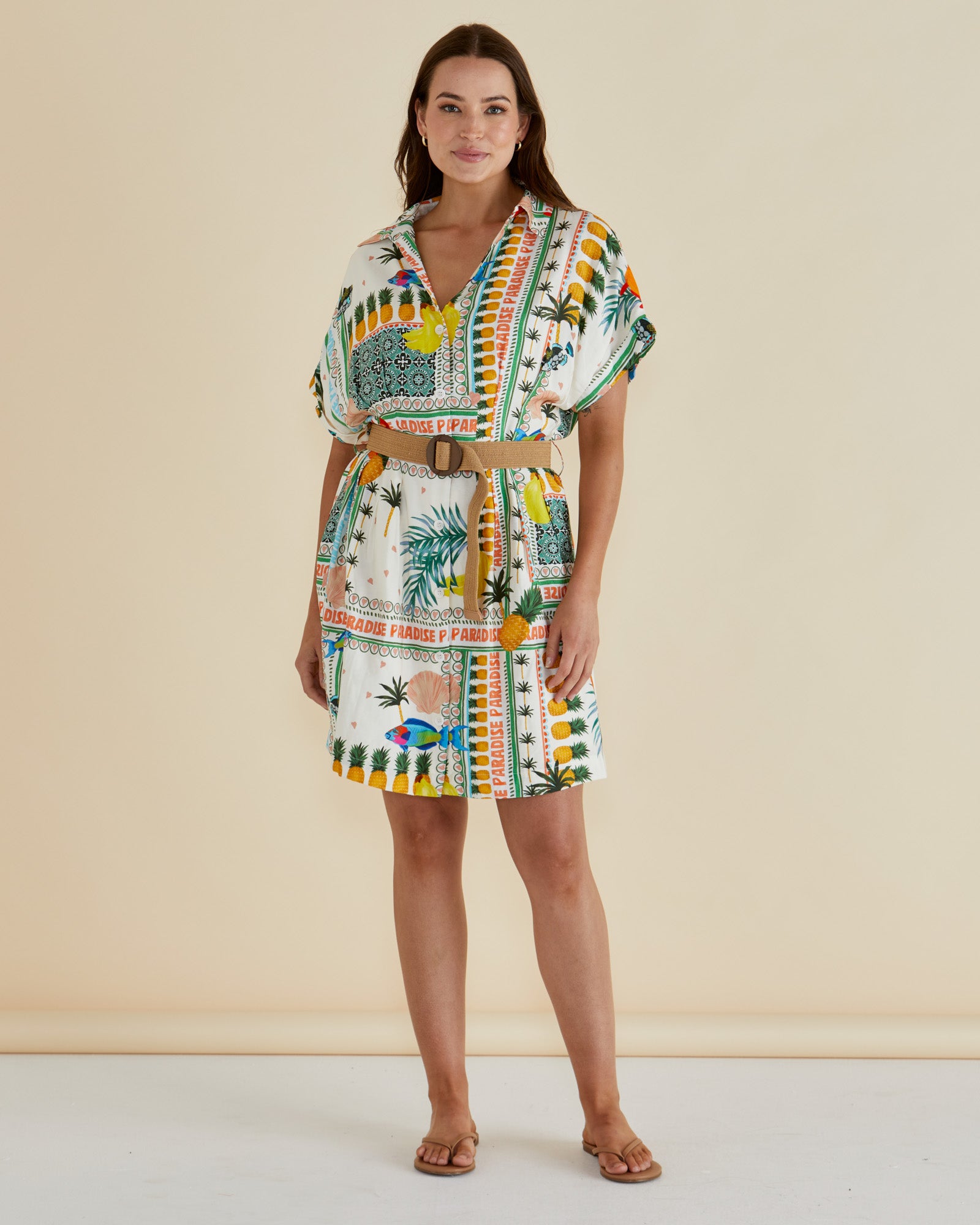 Woman wearing a colorful patterned belted mini shirt dress with paradise print with pineaples, leaves and fish on a white background with orange, green and blue print by betty basics.