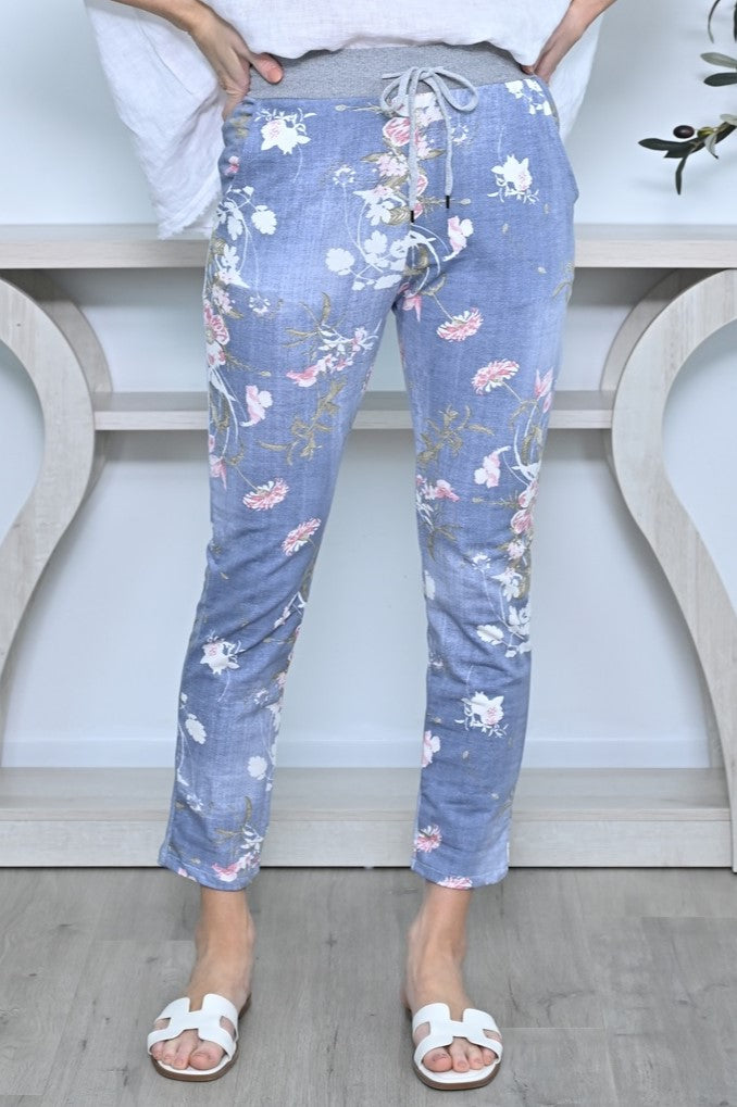 Lady wearing Cali&Co's rib waist stretch floral pants in denim blue (pale) floral print (in white and pink).