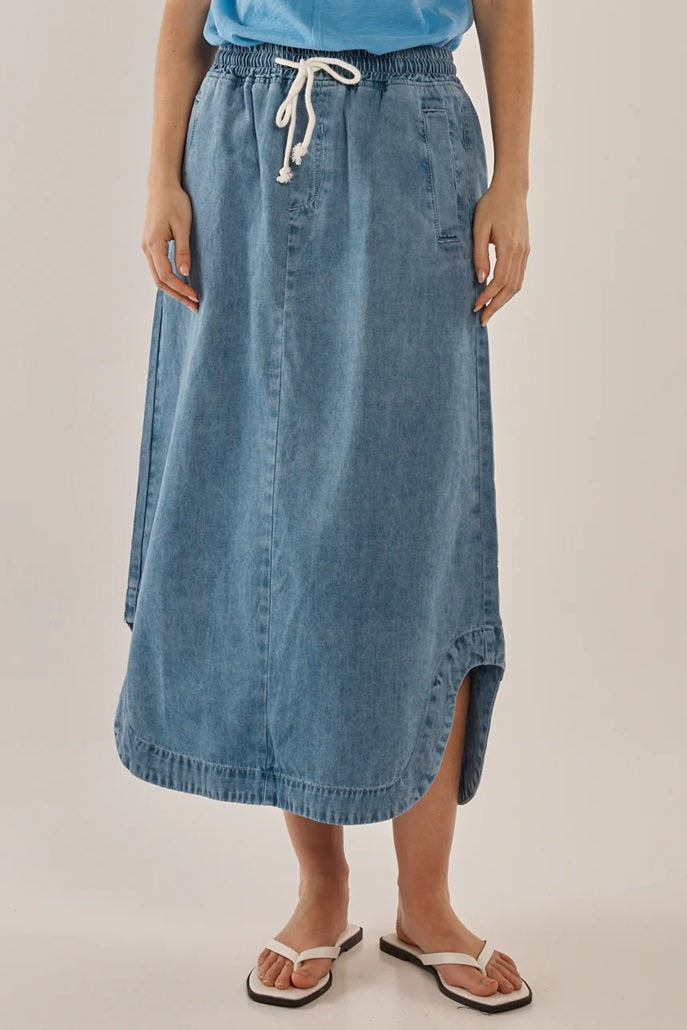 Woman wearing Betty Basic's Brunch Denim Skirt with a tie waist, pockets and a features curved side splits.