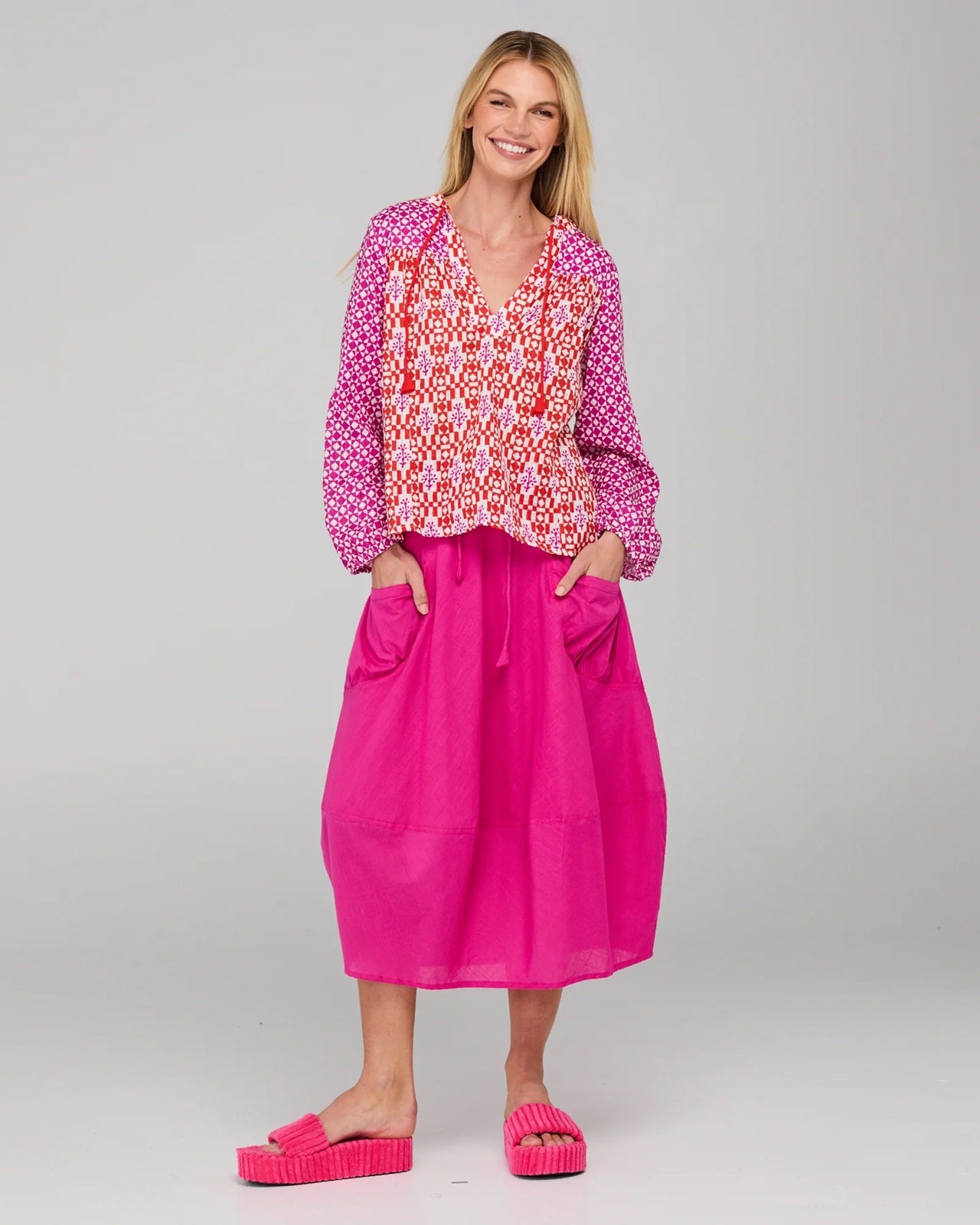 Model wearing a patterned cotton Fleetwood Top in Spliced Naples a pink, white and red print with long sleeves a v-neck and straight hem.