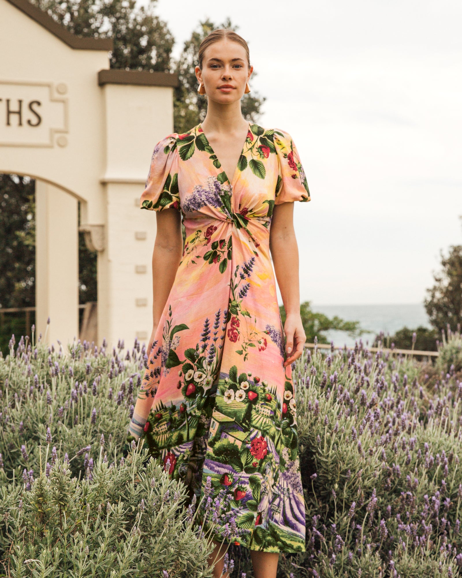 Model wearing the twist dress with strawberries and bulding print on a pink sunset background by white closet.